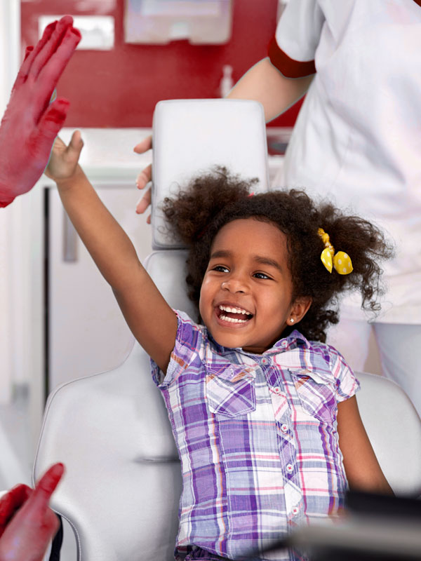 kinderbehandlung-2 Smiling young girl in a plaid shirt gives a high-five to a clinician in a dental clinic while seated in a chair.
