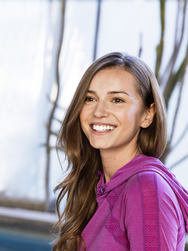 Zahnbegradigung Smiling woman with long light-brown hair wearing a purple hoodie, looking at the camera.